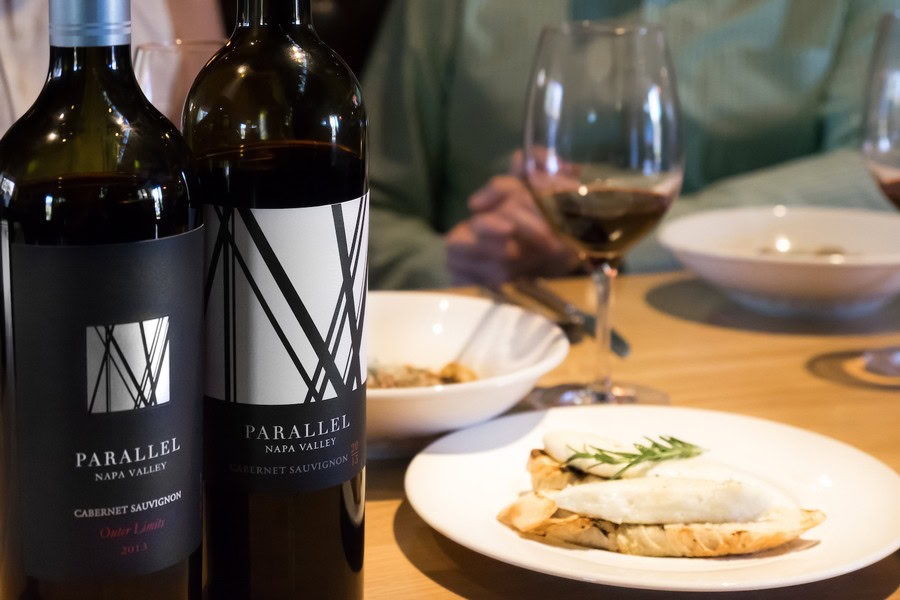 Two bottles of Parallel Wines Napa Valley Cabernet Sauvignon with a glass of red wine and a plate of food on a dining table.